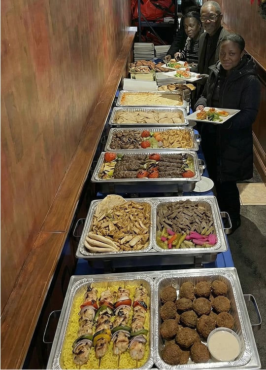Four people stand in line at a buffet table filled with trays of Mediterranean food, including skewers, pita bread, falafel, grilled meats, vegetables, and salad. The scene appears to be indoors.