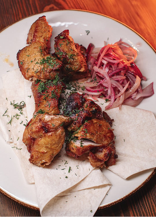 Grilled meat skewers served on flatbread, garnished with chopped herbs, accompanied by a side of marinated red onions on a white plate.