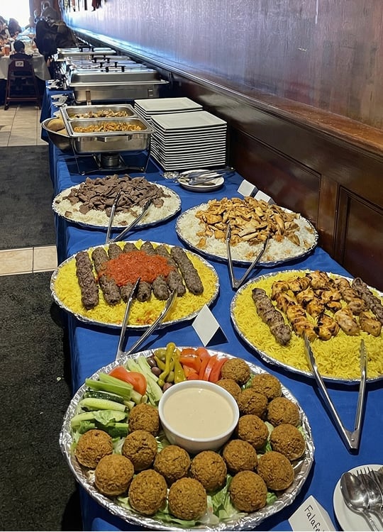 A buffet table with trays of Middle Eastern food, including falafel, kebabs, grilled chicken, beef, yellow rice, vegetables, and dipping sauce, with chafing dishes and stacks of plates in the background.