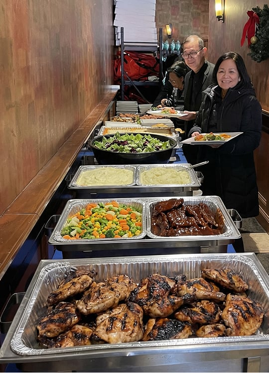 Three people serve themselves food from a buffet table filled with grilled chicken, vegetables, mashed potatoes, salads, and ribs in a warmly lit indoor setting.