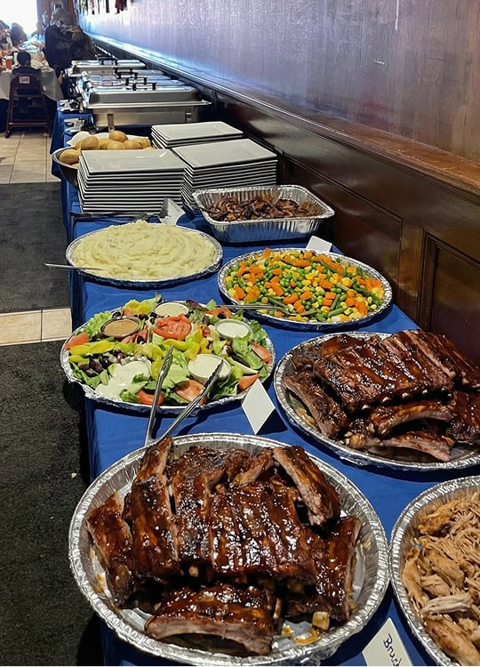 A buffet table features trays of glazed ribs, pulled pork, salad, mixed vegetables, mashed potatoes, bread rolls, and stacks of plates, all set against a wooden wall.