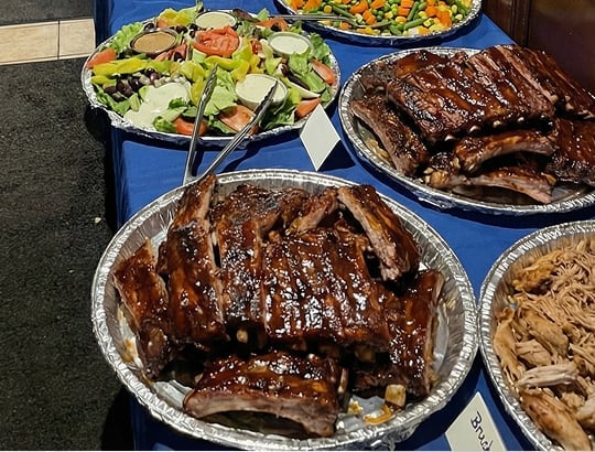Platters of glazed barbecue ribs, pulled pork, and mixed fresh salads with sliced vegetables are arranged on a blue tablecloth for a buffet-style meal. Serving tongs and labels are visible beside the dishes.
