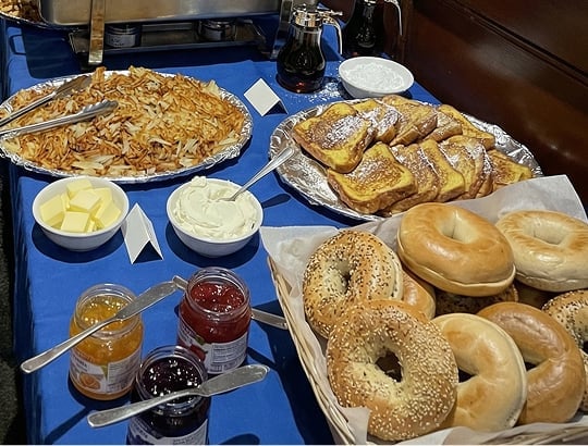 A breakfast buffet with hash browns, French toast, bagels, butter, cream cheese, syrup, and assorted fruit jams on a blue tablecloth.