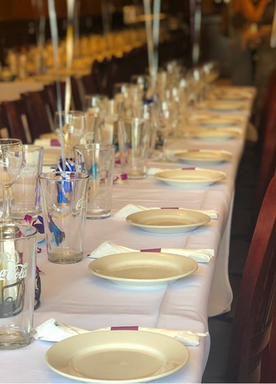 A long banquet table is set with white plates, glasses, and napkins on a white tablecloth. Balloons are tied along the table, and decorations include small purple and white flowers. The setting appears ready for a large gathering.