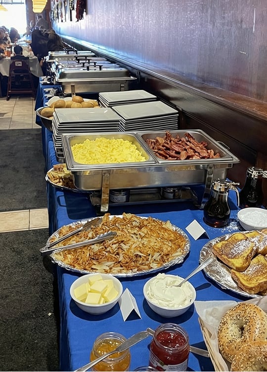 A breakfast buffet with plates, scrambled eggs, bacon, bread rolls, hash browns, bagels, bowls of butter, cream cheese, jams, and syrups on a blue tablecloth next to a wall. Diners are seated in the background.