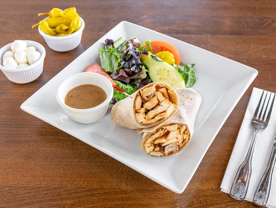 A square white plate with a chicken wrap cut in half, a small bowl of brown dipping sauce, and a fresh salad. On the side are bowls of pickled peppers and mozzarella balls, with a fork and knife on a napkin.
