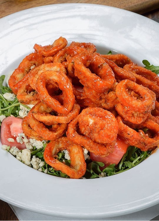 A white plate filled with a salad of arugula, diced tomatoes, and crumbled cheese, topped with crispy fried calamari coated in a reddish-orange sauce.