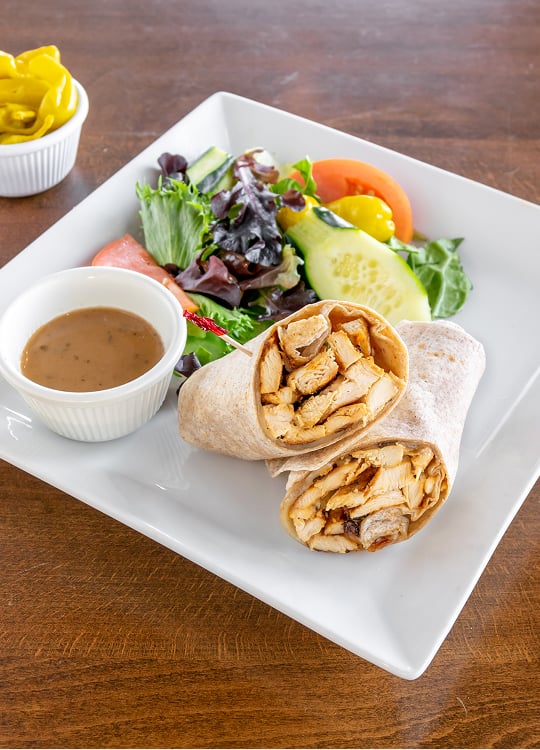 A plate with a sliced chicken wrap, a side salad with greens, cucumber, tomato, and pepperoncini, and a small cup of brown dressing. A small bowl of pepperoncini sits in the background on a wooden table.