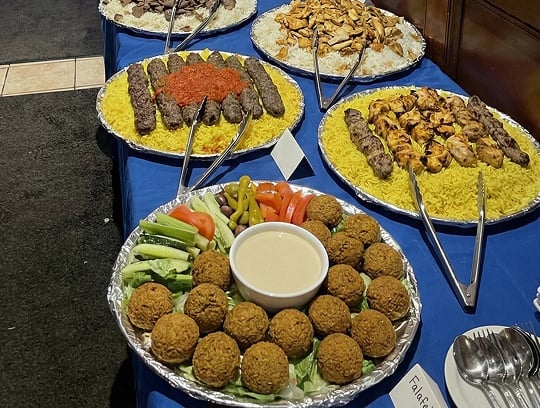 A buffet table displays platters of Middle Eastern food, including falafel with dipping sauce and vegetables, kebabs on yellow rice, and grilled chicken, all arranged on a blue tablecloth with serving tongs.