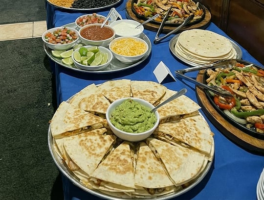 A buffet table with quesadillas arranged in a circle with guacamole in the center, surrounded by bowls of salsa, pico de gallo, cheese, sour cream, lime wedges, tortillas, and sautéed vegetables with chicken.