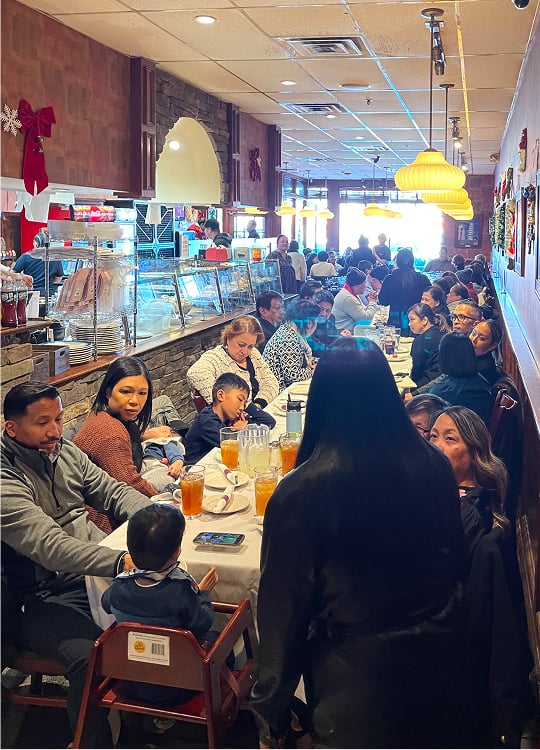 A crowded restaurant filled with people seated at tables, eating and chatting. Families and groups are gathered, with drinks on the tables and festive holiday decorations on the walls. The atmosphere is lively and warm.