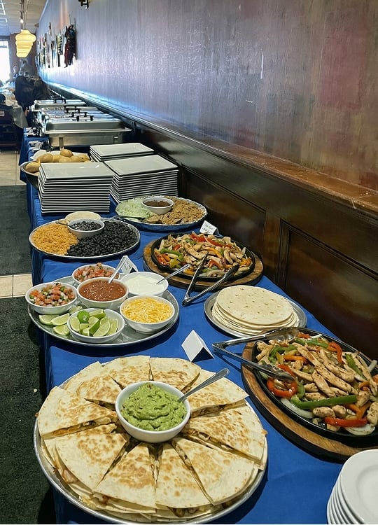 A buffet table with plates of quesadillas, grilled vegetables with chicken, tortillas, guacamole, shredded cheese, salsa, limes, and various toppings, all set on a blue tablecloth in a restaurant.