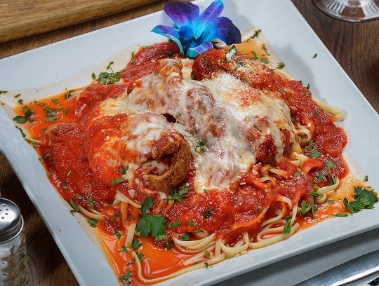 A plate of pasta topped with breaded, cheese-covered chicken, marinara sauce, and fresh parsley, garnished with a purple flower, is served on a square white dish.