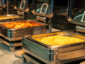 A buffet setup featuring trays of hot food, including a tray of yellow cooked noodles in the foreground and another tray with a mixed pasta dish in the background.