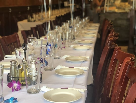 A long dining table set for a large group, with white tablecloth, plates, napkins, glasses, utensils, and scattered purple orchids. Wooden chairs line both sides of the table in a warmly lit restaurant.