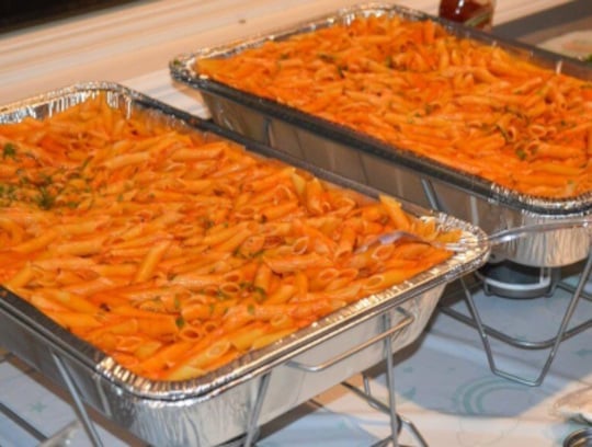 Two large aluminum trays filled with penne pasta in a red marinara sauce, garnished with green herbs, placed on wire racks for serving at an event or buffet.
