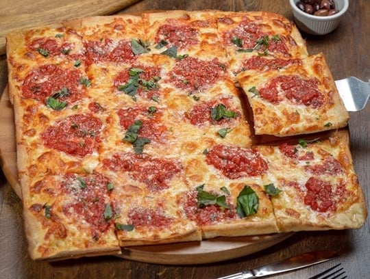 A rectangular pizza cut into squares sits on a wooden board. The pizza has melted cheese, tomato sauce dollops, and fresh basil leaves scattered on top. A metal spatula lifts one slice. A bowl of olives is in the background.