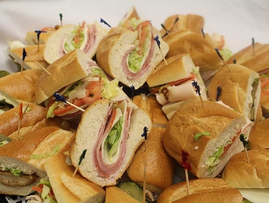 A platter of assorted sub sandwiches, each cut in half and held together with colorful toothpicks, filled with layers of deli meat, cheese, lettuce, and tomato on hoagie rolls.